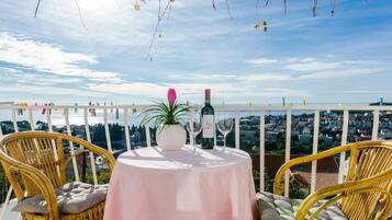 A balcony with wicker chairs, a table with a white tablecloth, a bottle of wine, and a vase with a flower.
