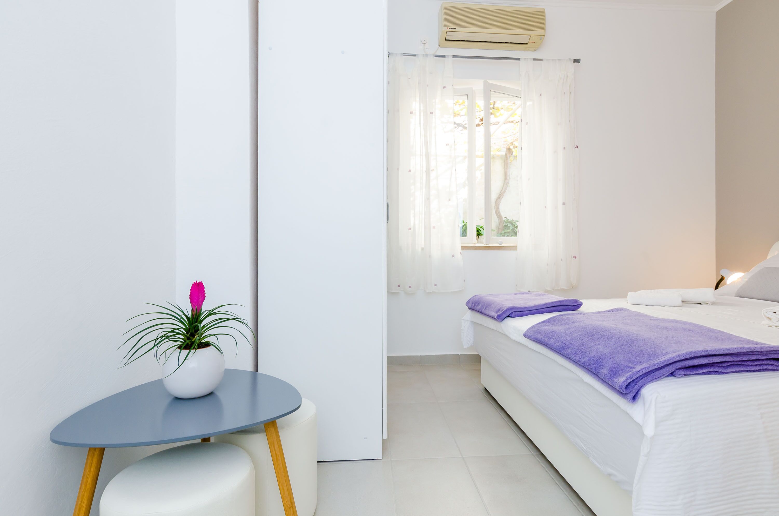 A hotel room with a bed, a small table with a potted plant, and a window with sheer curtains.