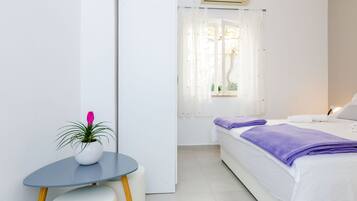 A hotel room with a bed, a small table with a potted plant, and a window with sheer curtains.