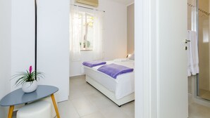 Un dormitorio con una cama, una mesita con una planta, un ventanal con cortinas y una zona de ducha.