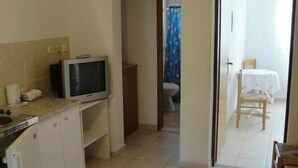 Cocina pequeña con televisor, teléfono y un armario. Se ve una puerta que da a un baño con inodoro y cortina de ducha. A través de una puerta abierta, se distingue una zona de comedor con mesa y sillas.
