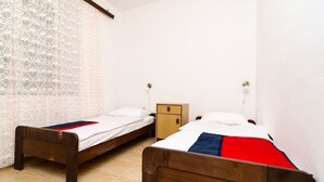 Dos camas de madera con ropa blanca y una colcha azul y roja, una mesita de noche de madera y una cortina estampada.