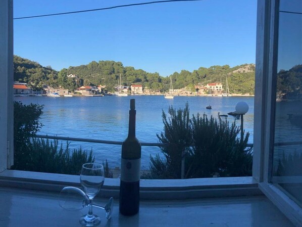 A bottle of wine and a glass on a windowsill with a view of a calm lake and buildings in the background.