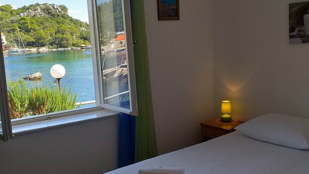 Ein Hotelzimmer mit Bett, Nachttisch mit Lampe, Fenster mit Blick auf einen See und Boote sowie einem Bild an der Wand.
