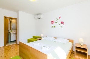 A bedroom with a wooden bed, green ottoman, white pillows, and a wall-mounted air conditioner.
