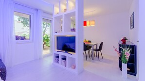 A modern living room with a TV, white shelving, a dining area, and a view of the outdoors. - Studio Luce Mala (ST) - Studio with Terrace (Split)