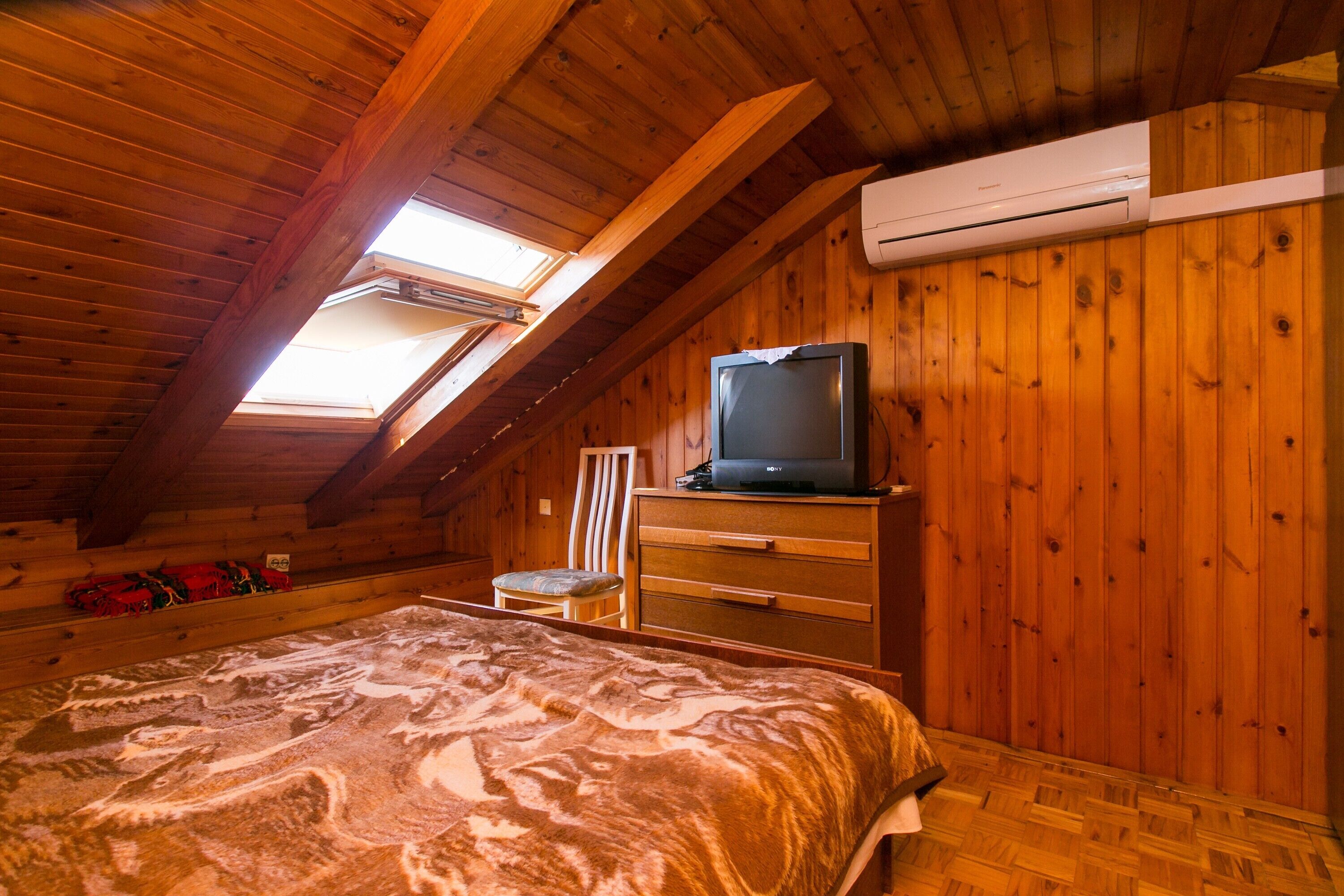 A cozy attic bedroom with a sloped ceiling, a bed with a patterned bedspread, a dresser, a television, and an air conditioning unit.