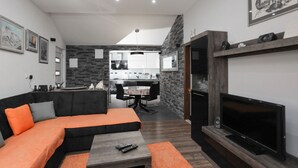 Un salon moderne avec un canapé noir et orange, un téléviseur et une cheminée. On aperçoit une cuisine à l’arrière-plan, avec des meubles blancs et une table à manger.