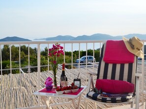 Un balcón con una mesa dispuesta con una botella, flores y fruta, una silla a rayas con un sombrero, y una vista de montañas y un coche al fondo.