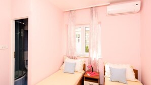 Une petite chambre à coucher décorée dans des tons roses, avec deux lits simples, une fenêtre avec des rideaux, un appareil de climatisation et un espace douche.