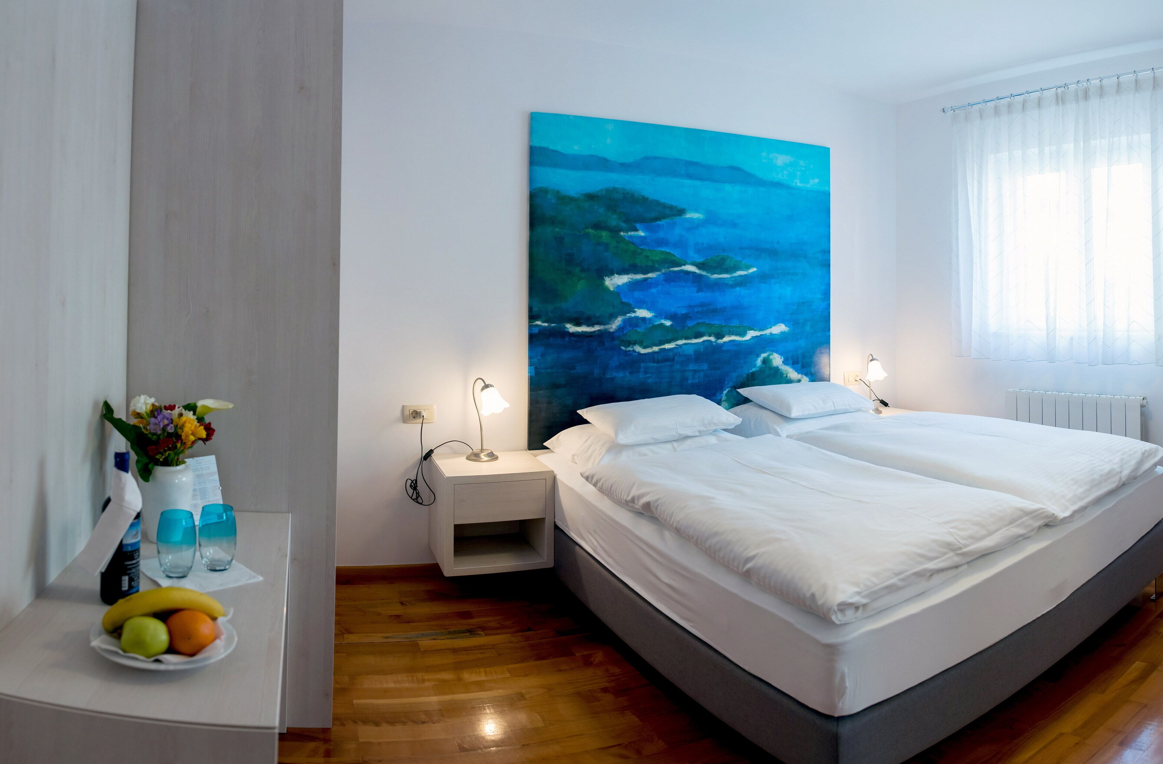 A modern hotel room with a large bed, a nightstand with a lamp, a fruit bowl, and a painting of a coastal scene.