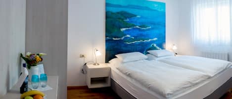 A modern hotel room with a large bed, a nightstand with a lamp, a fruit bowl, and a painting of a coastal scene.