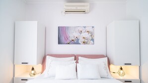 A bedroom with a bed, white walls, a pink headboard, two bedside tables with lamps, a wall-mounted air conditioner, and a floral painting above the bed.