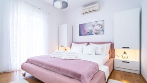 A modern bedroom with a large bed, white and pink bedding, a bedside lamp, and a wall-mounted air conditioner.