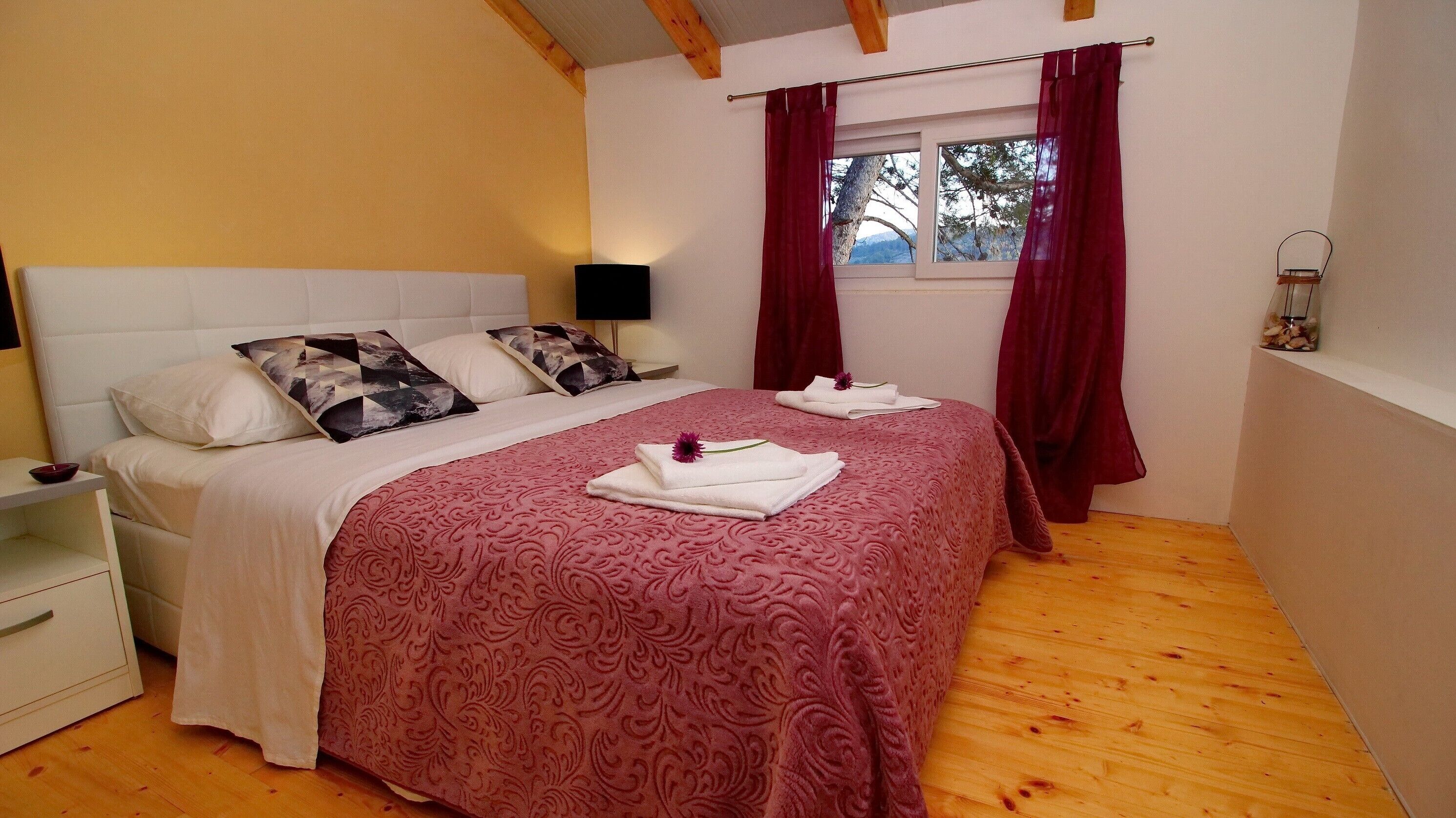 A bedroom with a double bed, a nightstand, a bedside table, a window with curtains, and a wooden floor.