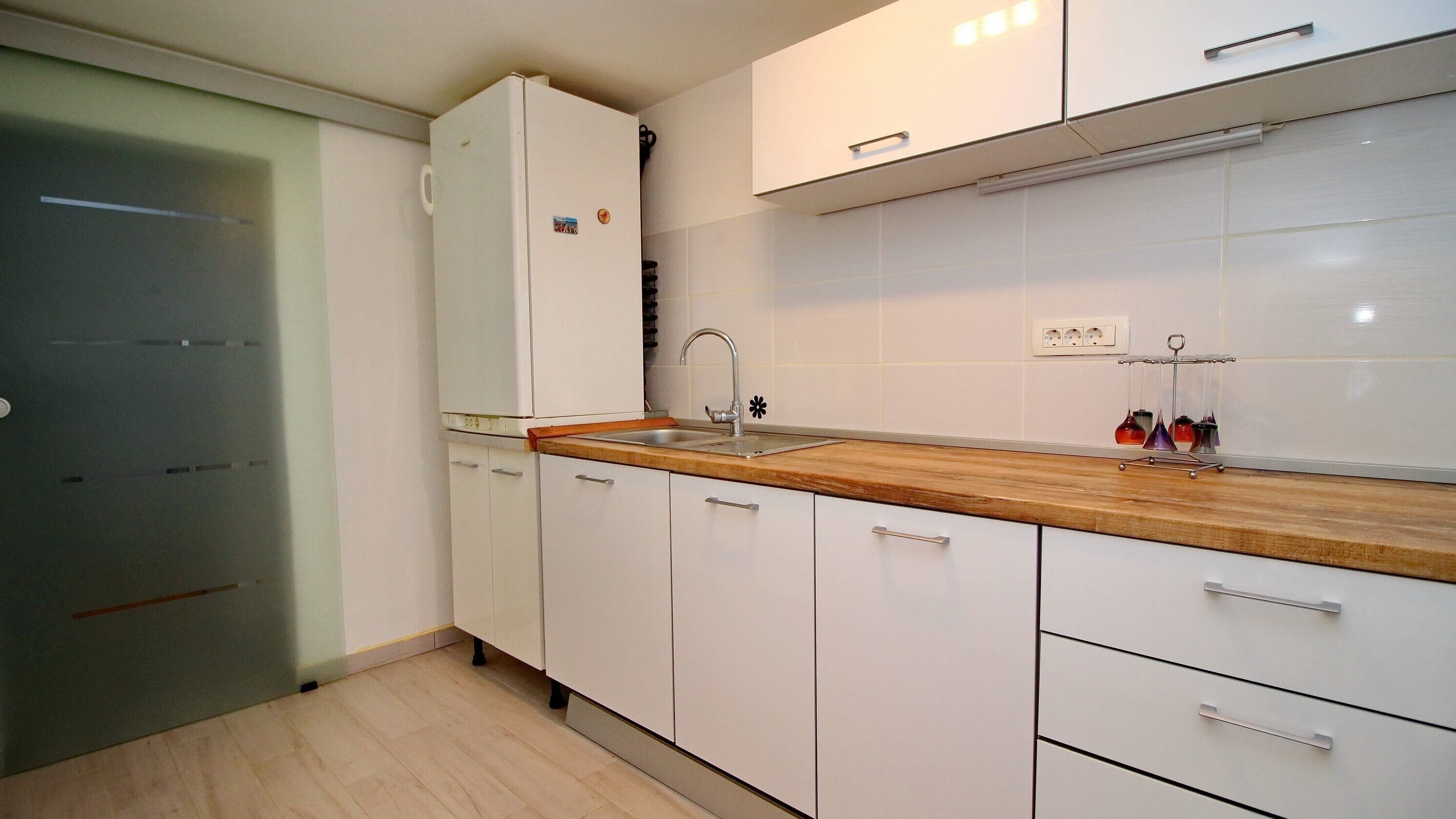 A kitchen with white cabinets, a wooden countertop, a stainless steel sink, and a refrigerator.