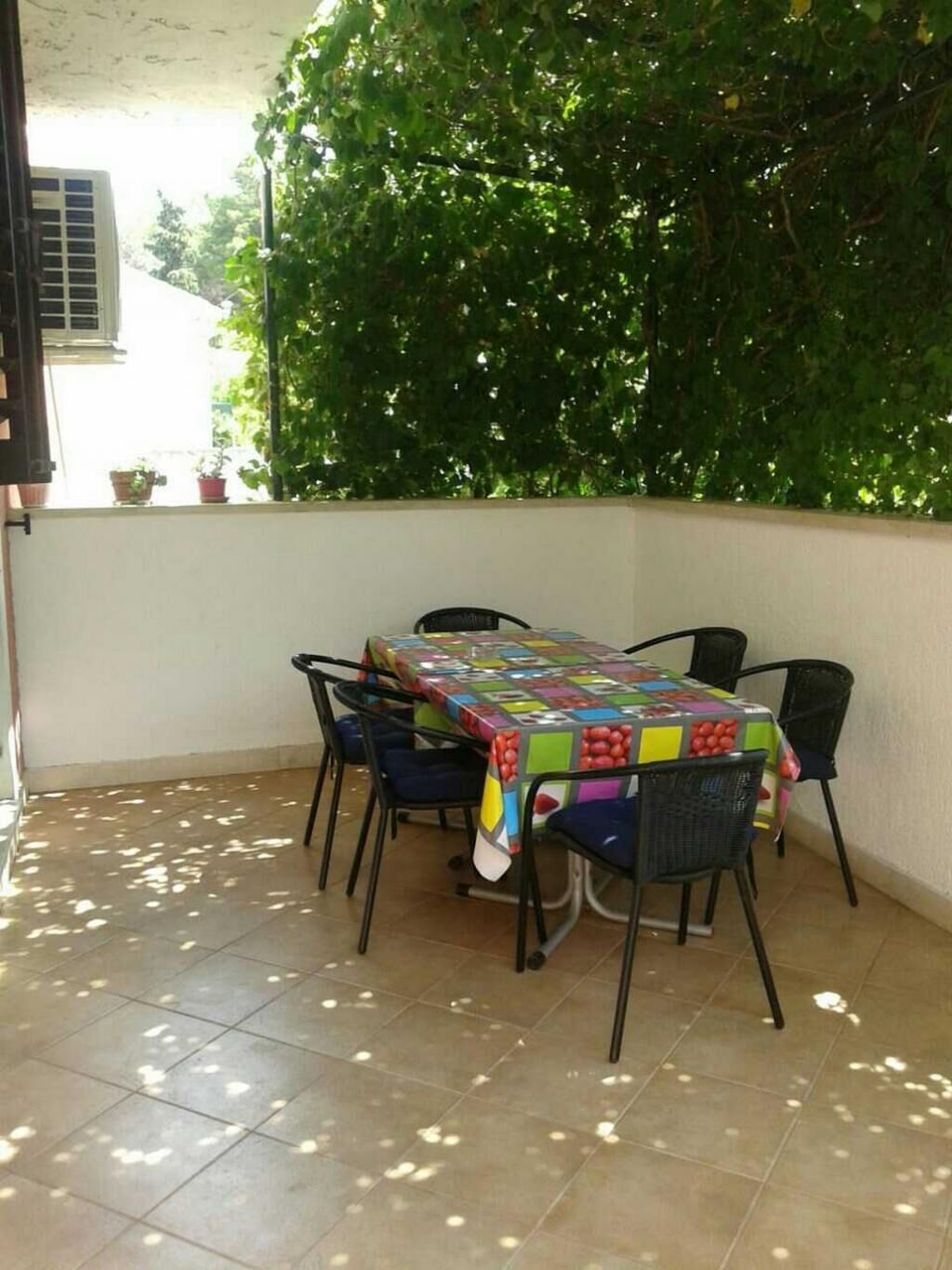 A covered patio with a table and chairs, surrounded by greenery and a white wall.