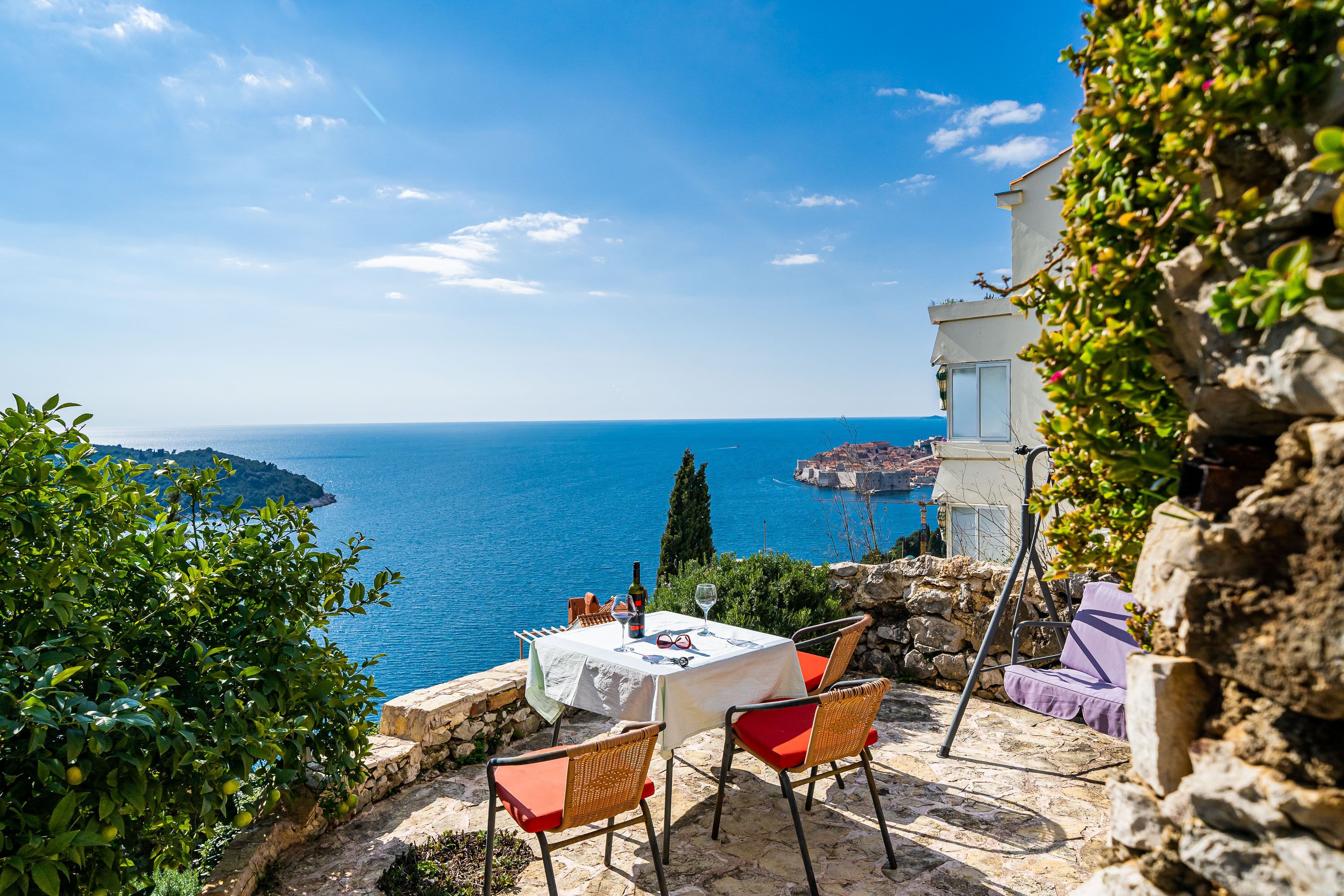 A table set with a white tablecloth, red cushions, and a bottle of wine, overlooking a coastal view.