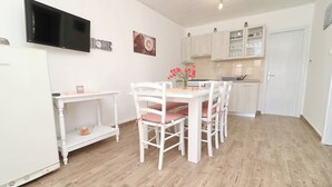 Een moderne keuken met witte kasten, een eettafel met stoelen, een aan de muur bevestigde televisie en een klein plankje met een kaars.