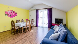 A dining area with a table and chairs, a blue sofa, and a TV on a stand. - Apartments Mali Paradiso - Two Bedroom Apartment with Balcony and Pool View (Mlini)