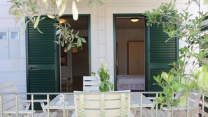Un patio con mobiliario blanco y contraventanas verdes, una mesa blanca con un florero y vista al interior del edificio.