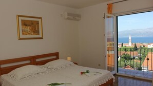 Un dormitorio con cama de madera, colcha de encaje blanco, un cuadro en la pared y un balcón con vistas a un pueblo y al mar.