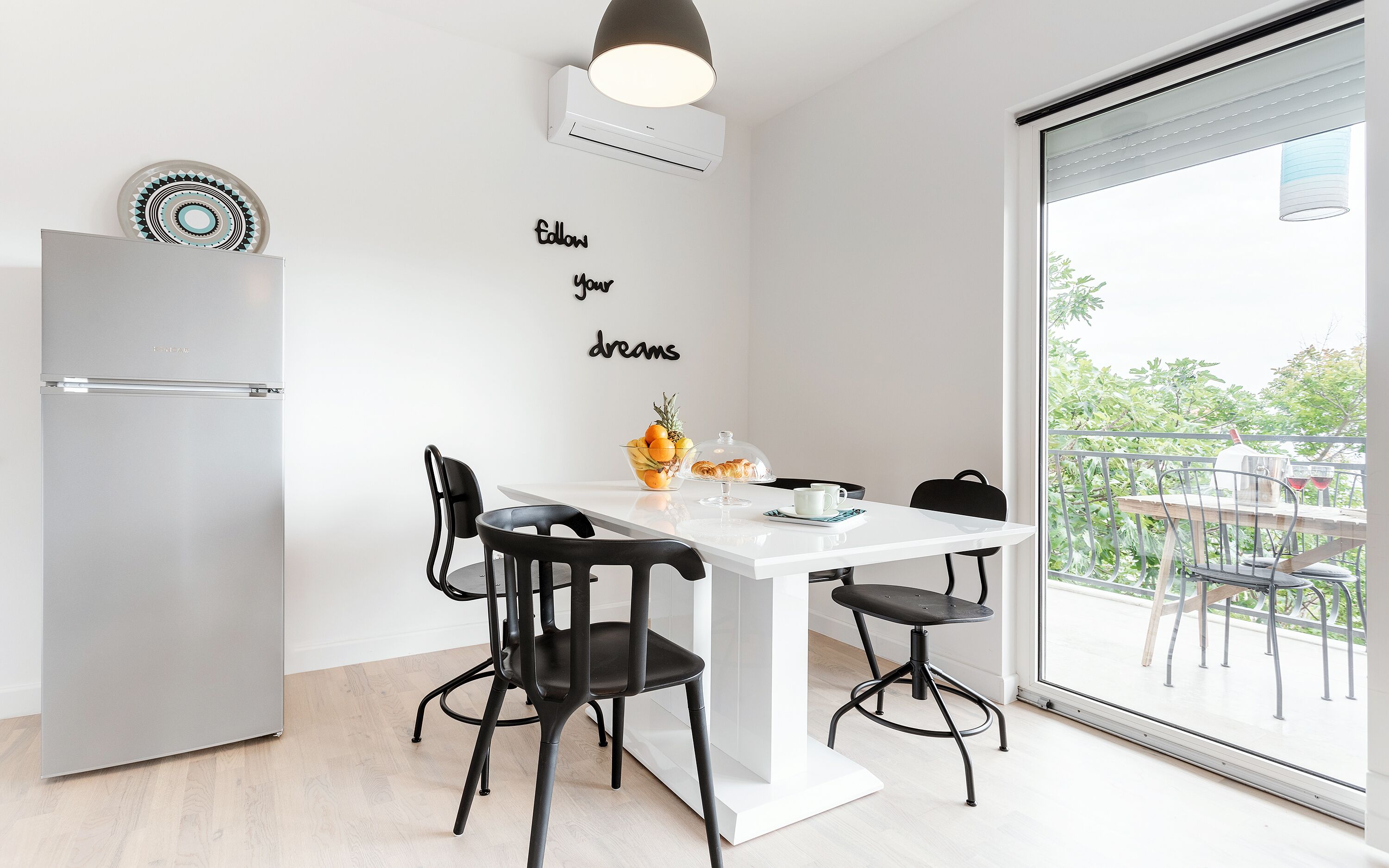 Une cuisine moderne avec un réfrigérateur blanc, une table à manger entourée de chaises noires et une vue sur la verdure grâce à une grande fenêtre.