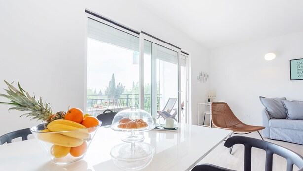 Ein moderner Essbereich mit einem weißen Tisch, transparenten Schalen voller Obst und Blick ins Freie durch große Fenster.