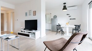 Ein modernes Wohnzimmer mit Flachbildfernseher, einer Essecke mit weißem Tisch und Stühlen sowie einer Küche mit Kühlschrank.