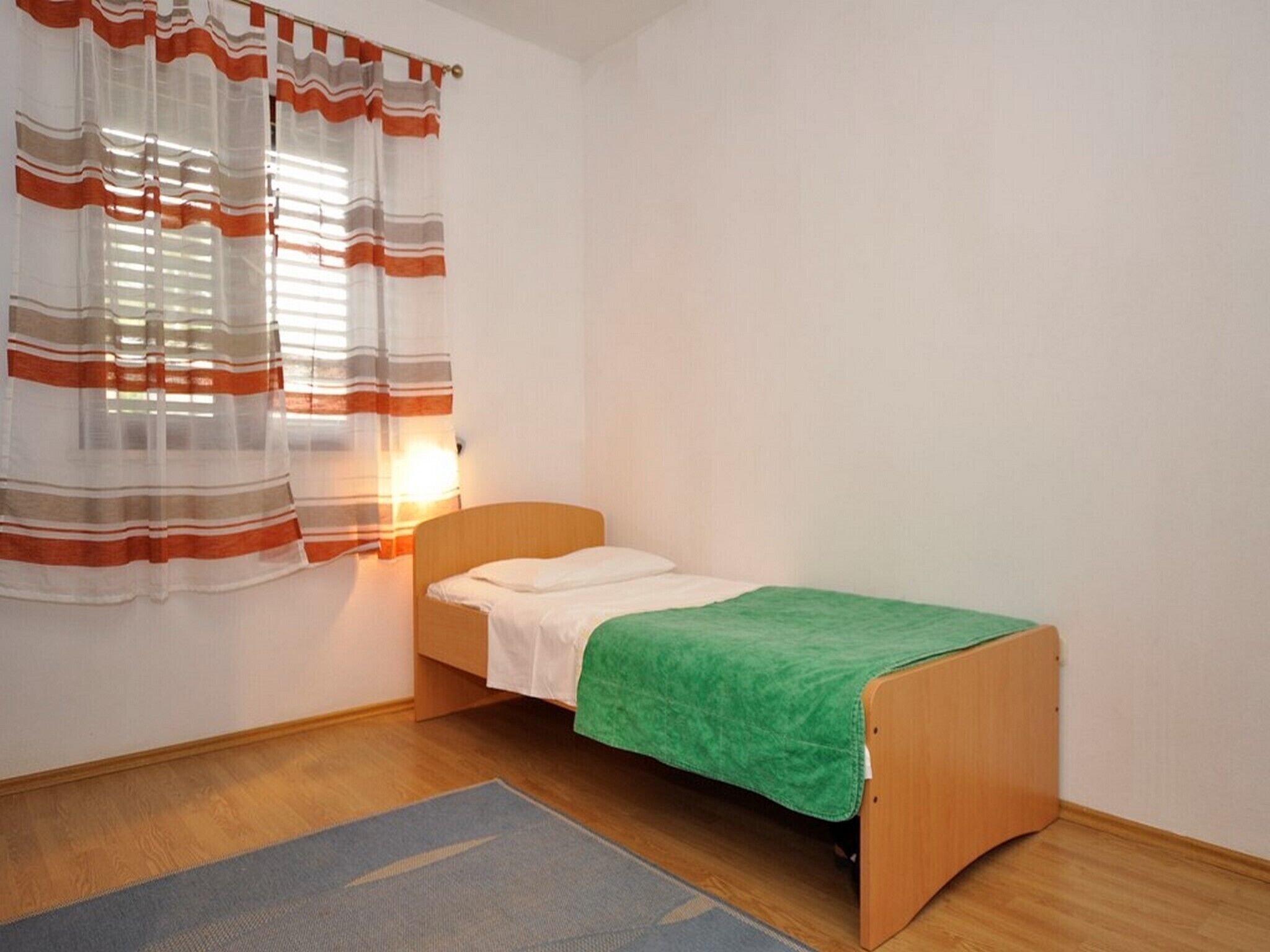 Une petite chambre avec un lit simple, une tête de lit en bois, une couverture verte et un tapis bleu.