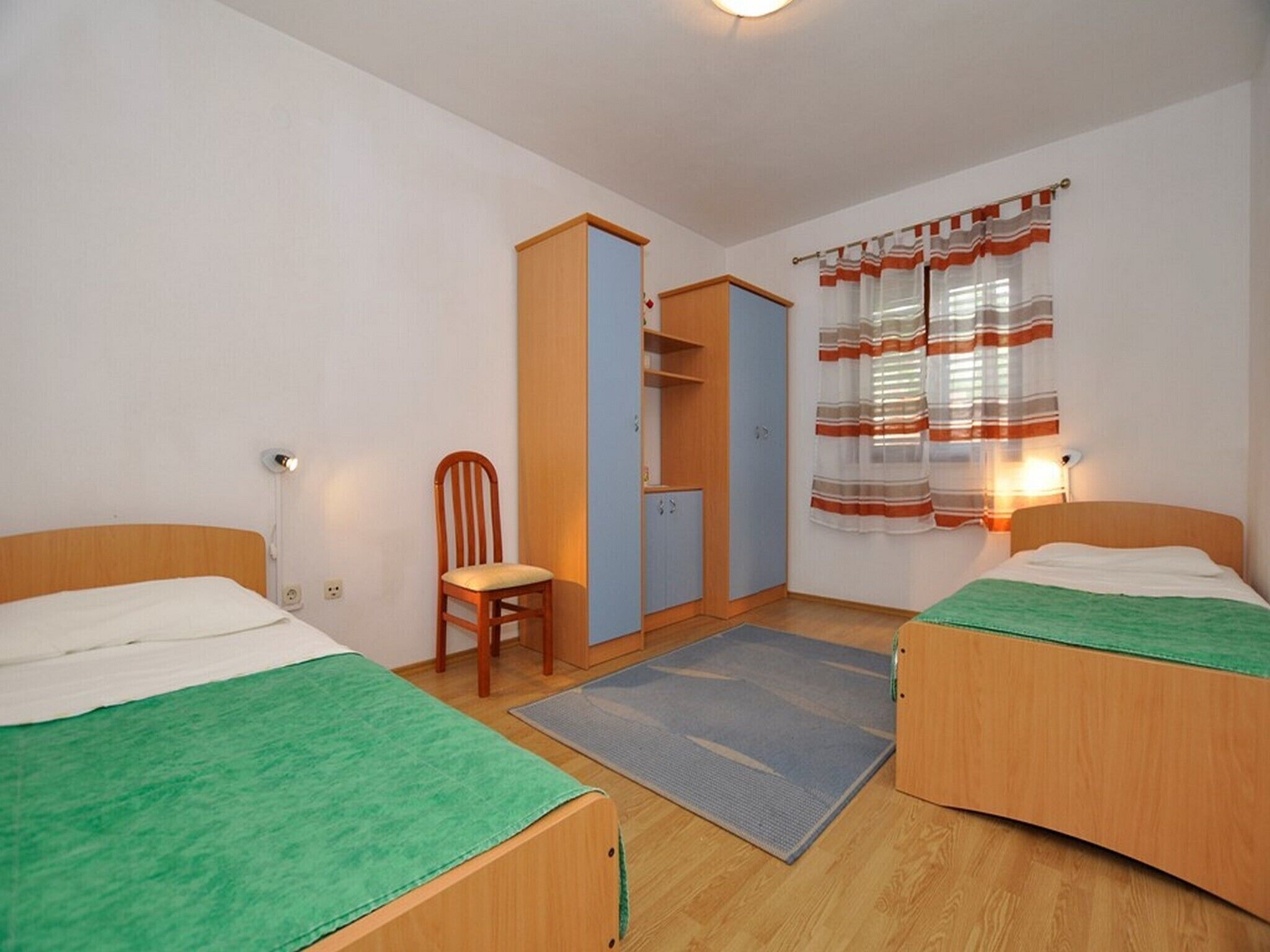 Une petite chambre propre, avec deux lits simples, une chaise, une armoire et une fenêtre avec des rideaux.