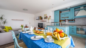 Ein modernes Wohnzimmer mit einer Couch, einem Esstisch gedeckt mit Frühstück und einer Küche mit türkisfarbenen Schränken.