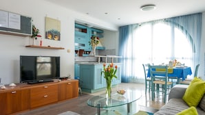 Ein modernes Wohnzimmer mit Fernseher, Sofa, Glastisch und einem Essbereich mit blauer Tischdecke.