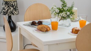 Zona pranzo con tavolo bianco, due sedie, un orologio, una lampada, un vaso con fiori e prodotti per la colazione, tra cui croissant e succo d'arancia.