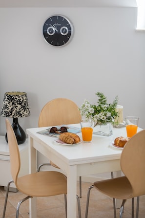A dining area with a white table, two chairs, a clock, a lamp, a vase with flowers, a candle, and breakfast items including croissants and orange juice.