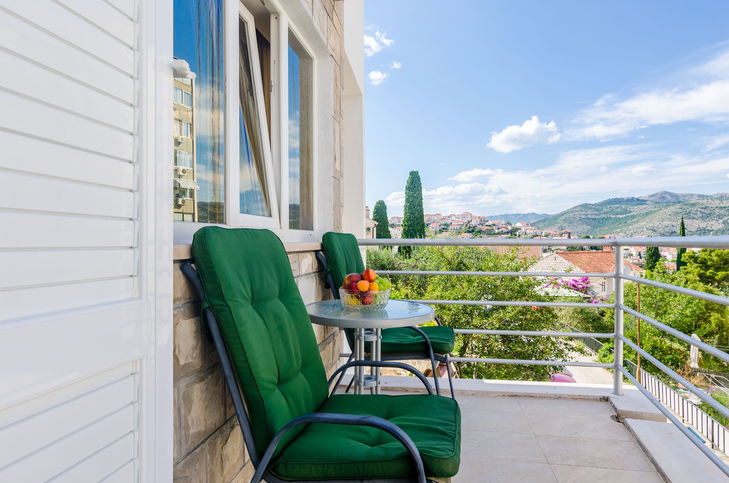 A balcony with green cushioned chairs and a table with a fruit bowl.
