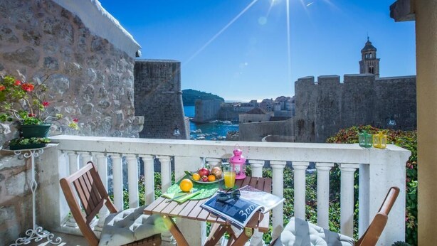 A balcony with a table set for a meal, overlooking a historic cityscape with a clear blue sky and a bright sun.