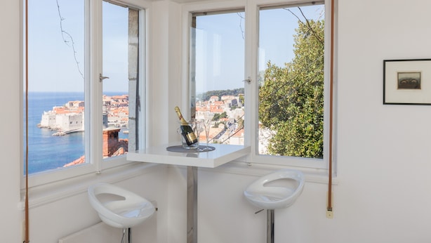 A corner room with two white bar stools, a small table with a bottle and glasses, and a view of a coastal town through large windows. - Apartment Rea-Three Bedroom Apartment with Sea View (Dubrovnik)