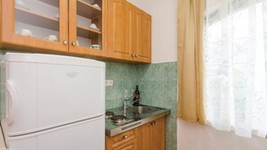 A kitchen with wooden cabinets, a white refrigerator, a sink with a wine bottle, and a window with patterned curtains.