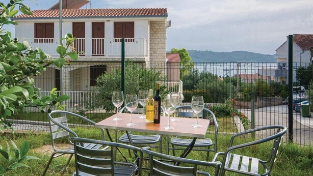 A dining table with chairs and wine glasses, set against a scenic backdrop of a house and a body of water.