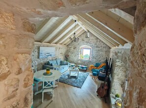 Un intérieur rustique en pierre, avec un plafond haut, un mur en pierre, un canapé, une table, une guitare et une fenêtre.