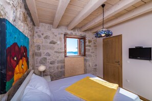 Een slaapkamer met een bed, een schilderij, een houten plafond, een stenen muur, een houten deur en een televisie.