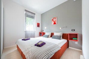 Ein modernes Schlafzimmer mit einem großen Bett, einem Fenster mit Jalousien, einem roten Regal und einer Wandlampe.