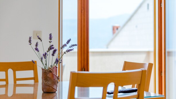 Ein Esstisch mit einer Vase voller Lavendelblumen, Holzstühle und Blick auf Gebäude durch große Fenster.