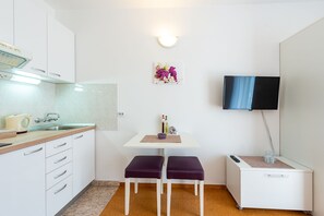 Una cocina compacta con armarios blancos, una encimera, un lavabo y una pequeña mesa con dos taburetes morados. Se observa también un televisor de pared y un accesorio de iluminación.