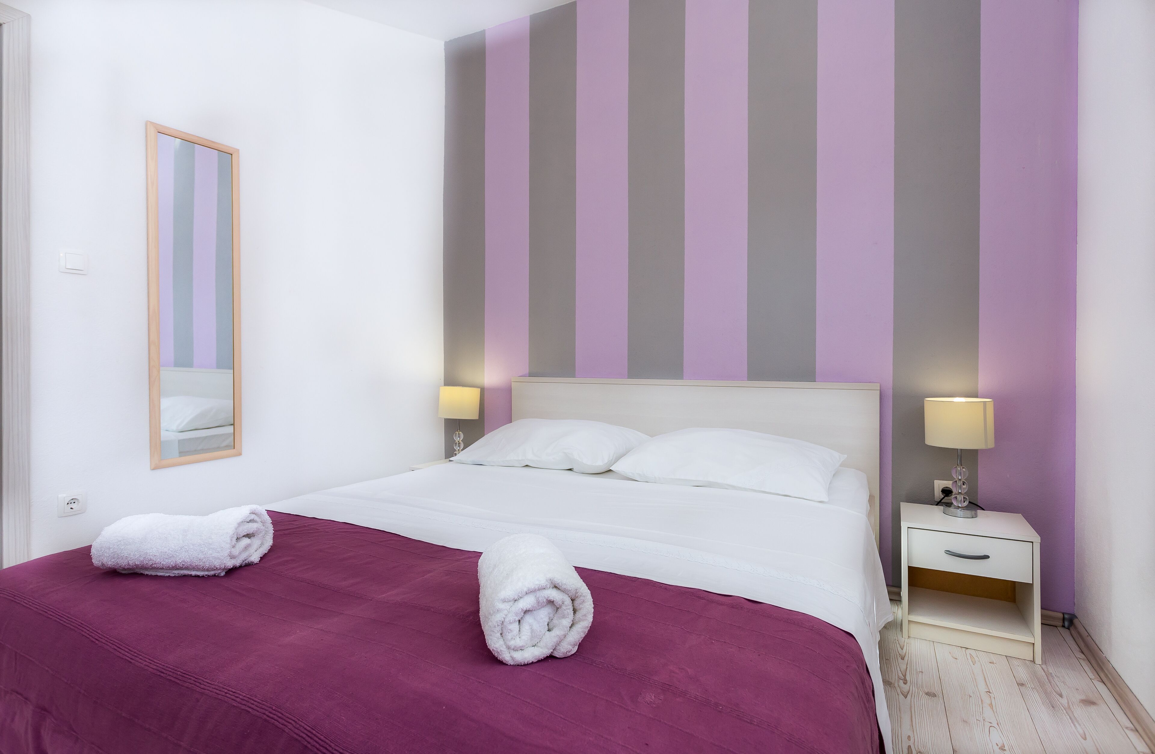 Ein ordentlich bezogenes Bett mit weißen Leinen, einer violetten Tagesdecke und zwei aufgerollten Handtüchern. Ein Nachttisch mit Lampe, und ein Spiegel sind an der Wand angebracht.