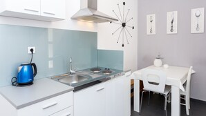 A modern kitchen with white cabinets, a blue kettle, a gas stove, and a dining area with a white table and chairs. - Magdales - Two Bedroom Apartment with Balcony (Omis)