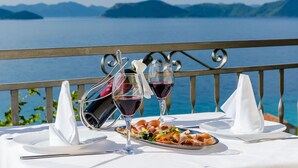 Une table dressée avec une assiette de nourriture, deux verres à vin et une bouteille de vin, le tout placé sur un balcon donnant sur une étendue d’eau.