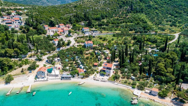 Vue aérienne d’un village côtier avec des maisons, une plage et des eaux turquoise et cristallines.