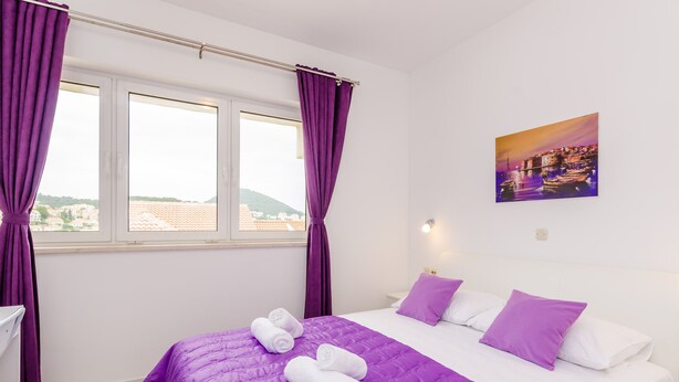 Ett hotellrum med en säng, lila gardiner, en tavla och utsikt över byggnader.